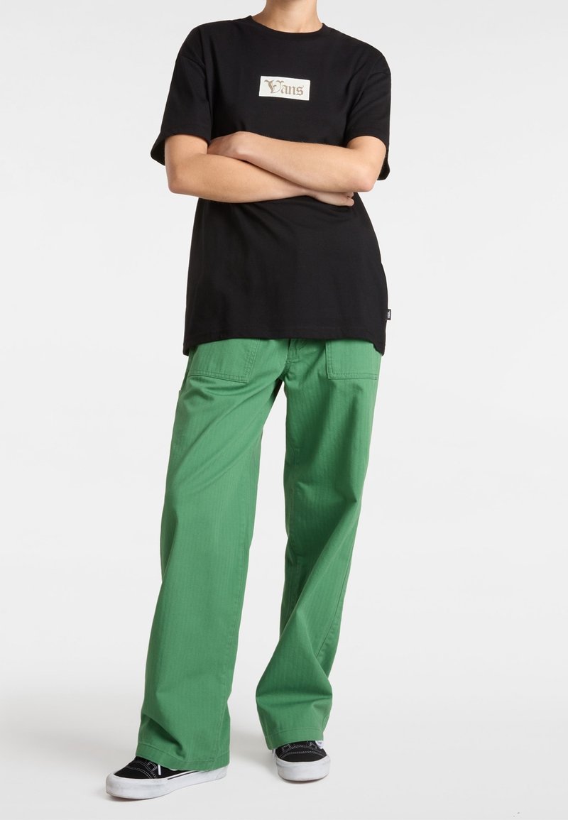 Sort bomulds t-shirt med et hvidt Vans-logo, parret med grønne vide bukser. Outfittet afsluttes med sort-hvide sneakers.