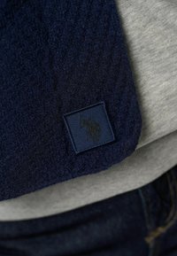 Marineblå fleecestof med en struktureret overflade og et kvadratisk patch med et mørkt logo. Grå stof synligt nedenunder. Denim synligt nedenfor.