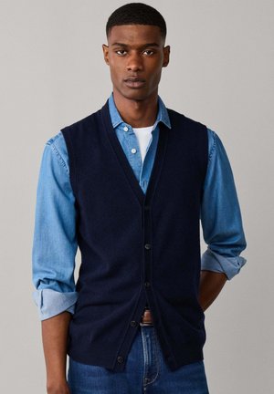 Jonge man met een marineblauwe, geknoopte vest over een lichtblauw opgerold denimshirt en blauwe jeans, staand tegen een effen achtergrond.