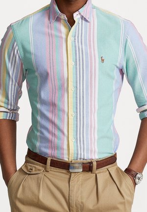 Homme portant une chemise boutonnée à rayures verticales multicolores pastel avec un pantalon kaki, une ceinture marron et une montre-bracelet.