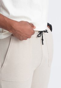 Pantalones texturizados beige con cintura ajustable, que presentan un patrón en blanco y negro. Primer plano de una mano descansando sobre la cinturilla.