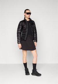 Sort pufferjakke med quiltet tekstur, parret med en sort kjole. Sorte sneakers og sokker fuldender outfittet. Modellen står imod en neutral baggrund.
