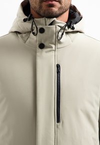 Lichtbeige jas met een hoge kraag en een verstelbare capuchon. Bevat een ritsborstzak, zwarte accenten en een slanke, gladde stof.