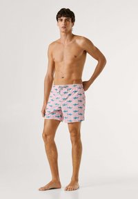 Mladý muž stojící naboso, oblečený v růžových plavkách s modrými potisky žraloků, jednou rukou za zády, neutrální výraz.