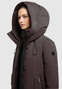 Nafukovacia hnedá bunda s veľkou kapucňou, vybavená zipsovým zapínaním, šnúrkami a nášivkou na rukáve. Hladký povrch a polstrovaný dizajn.
