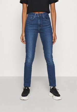 Personne portant un jean slim bleu taille haute et des baskets noires et blanches, debout contre un mur et un sol gris uni.