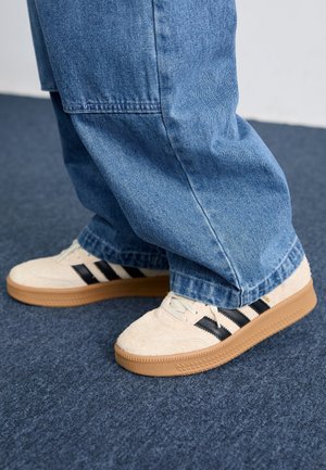 Osoba nosząca jasnobeżowe buty Adidas z czarnymi paskami i gumowymi podeszwami, w połączeniu z luźnymi niebieskimi dżinsami, stojąca na ciemnym dywanie.