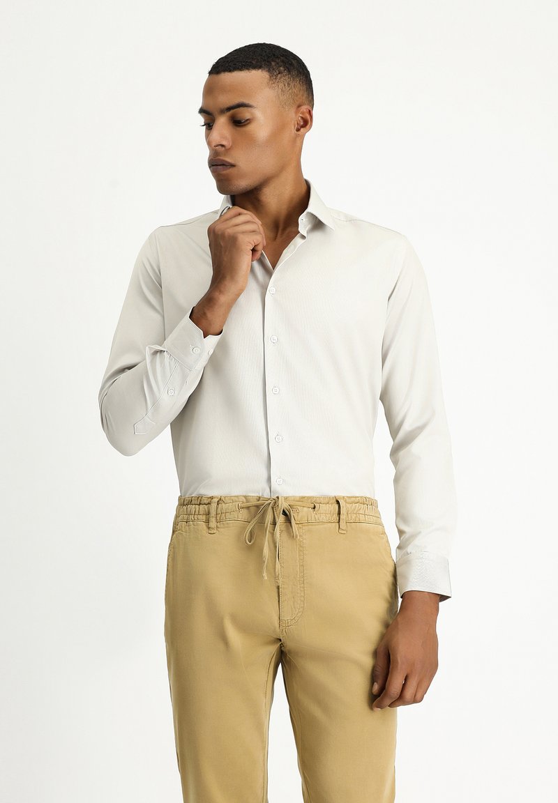 Camicia button-up grigio chiaro con colletto strutturato, maniche lunghe e una sottile trama, abbinata a pantaloni beige con coulisse.