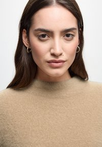 Beige, fluffig tröja med hög halsringning, med en mjuk textur. Modellen bär guldiga ringörhängen och har långt, mörkt hår.