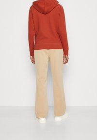 Persoon in een roestkleurige hoodie, beige wijde broek en witte sneakers staat met de rug naar de camera tegen een eenvoudige witte achtergrond.