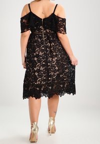 Schwarzes Spitzenkleid mit schulterfreiem Design, floralem Muster, Reißverschluss auf der Rückseite und knielangem Saum; kombiniert mit metallisch goldenen Absatzschuhen.