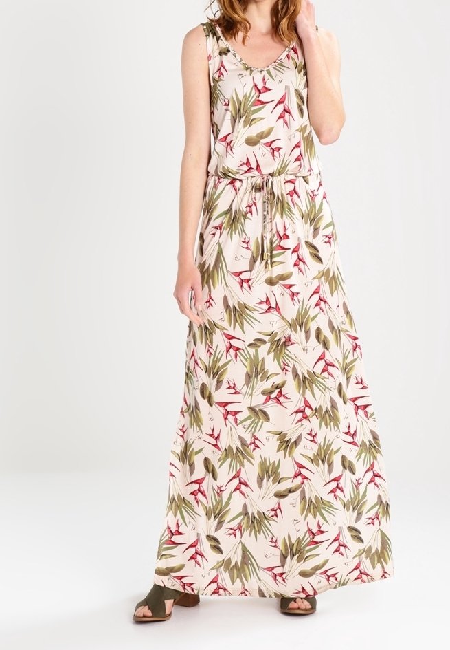 Femme portant une robe fleurie sans manches, longue jusqu'au sol, avec des feuilles vertes, rouges et beige, associée à des sandales vertes à bout ouvert, debout sur fond blanc.