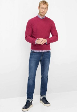 Mann steht mit verschränkten Händen, trägt einen roten Pullover über einem geblümten Hemd, blaue Jeans und hochgekrempelte schwarze Sneakers.