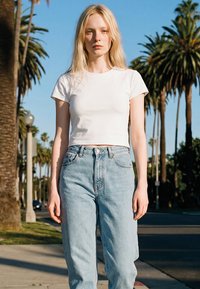 Mujer rubia con camiseta blanca recortada y vaqueros de tiro alto azul claro, de pie en la calle con palmeras al fondo.