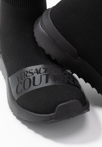 Black knit slip-on sneakers featuring a textured sole and a glossy black band with embossed "VERSACE JEANS COUTURE" logo across the midfoot.