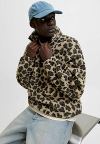 Leopardmønstret fleecejakke med høj krave, frontlynlås og en blød tekstur. Kombineret med en lyseblå cap og lyse jeans.