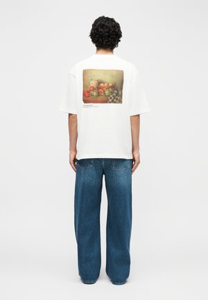 Άνδρας με σγουρά σκούρα μαλλιά, φοράει λευκό φαρδύ T-shirt με εκτύπωση φρούτων και χαλαρό μπλε τζιν, στέκεται με την πλάτη προς το λευκό φόντο.