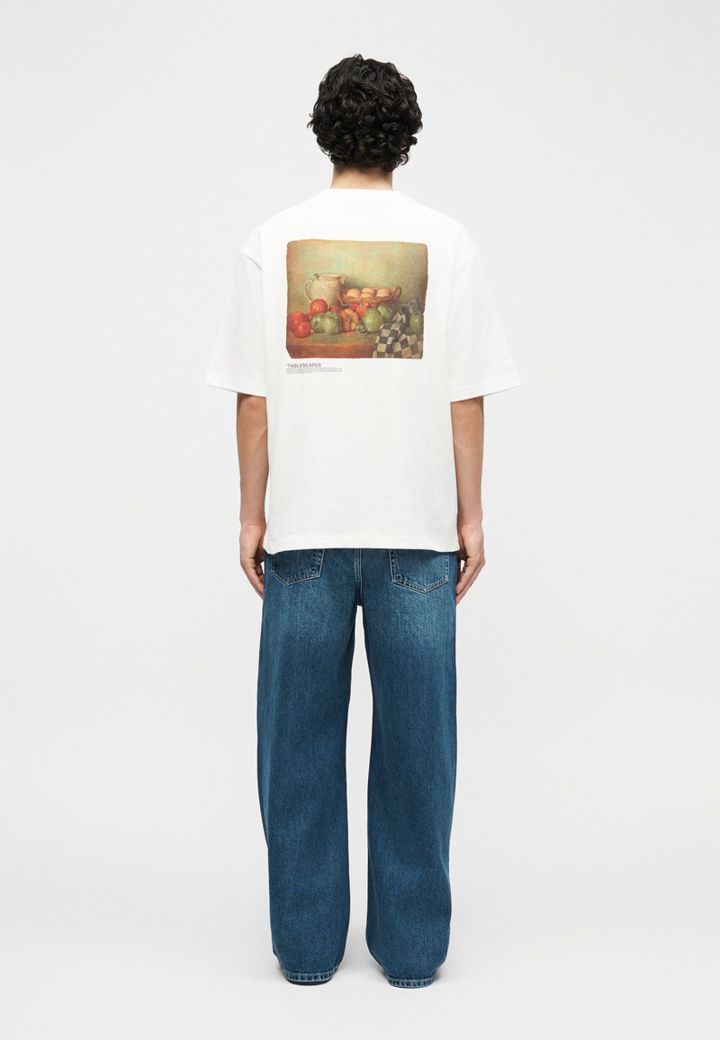 Άνδρας με σγουρά σκούρα μαλλιά, φοράει λευκό φαρδύ T-shirt με εκτύπωση φρούτων και χαλαρό μπλε τζιν, στέκεται με την πλάτη προς το λευκό φόντο.