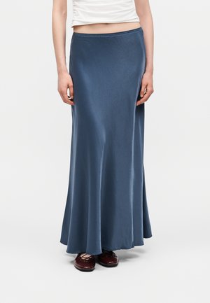 Žena nosi dlhú, prúdiacu modrú sukňu, biely krátky top a bordové baletky, stojí na jednoduchom bielom pozadí.