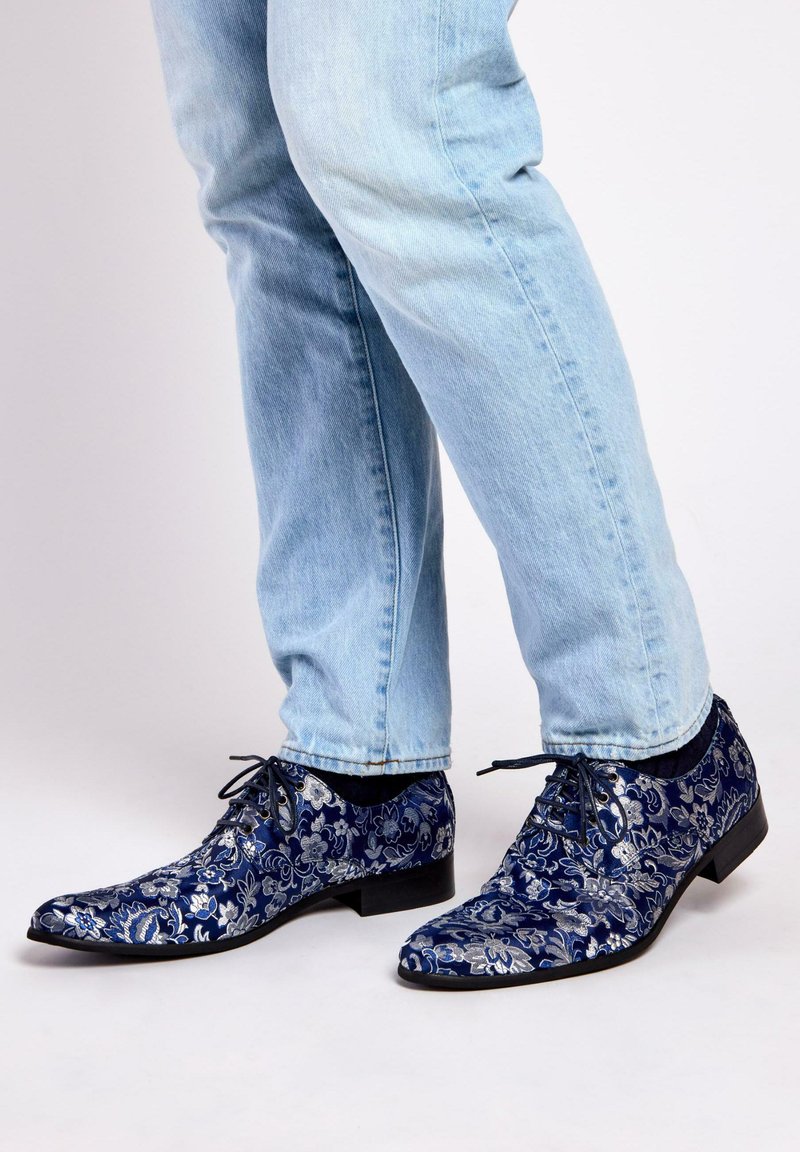 Navy blue dress shoes with floral silver pattern, pointed toe, and lace-up design. Worn with light blue jeans, showcasing patterned detail.