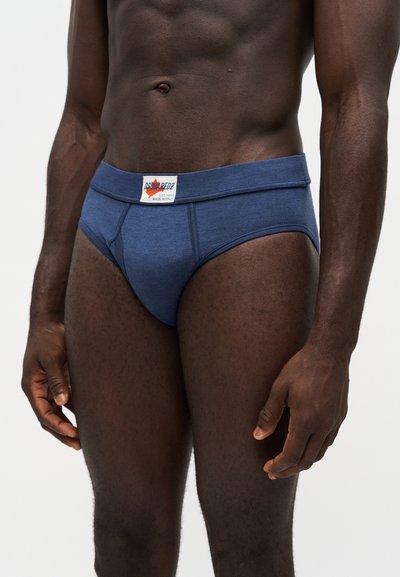 Modèle masculin portant un slip bleu Dsquared2 avec un écusson logo sur la ceinture, debout devant un fond clair uni.