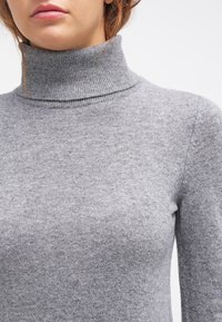 Kvinna i en grå stickad polotröja, som visar nacken och övre delen av överkroppen mot en ljus bakgrund.