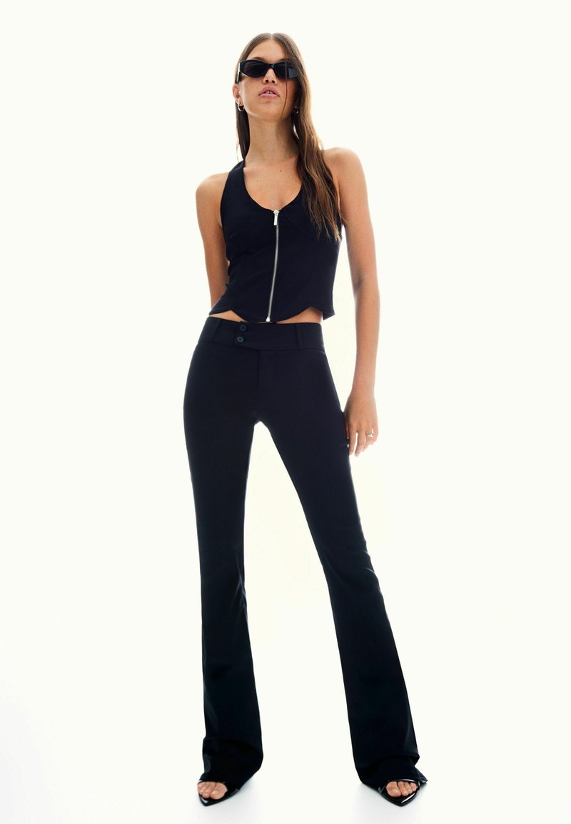 Zwarte cropped tanktop met een rits aan de voorkant, gecombineerd met hooggetailleerde zwarte flared broeken. Model draagt donkere zonnebrillen en hoge hakken sandalen.