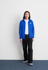 Jaqueta azul com zíper e detalhes brancos, usada sobre uma t-shirt branca. Calças pretas e ténis brancos completam o visual, em pé sobre um tapete.