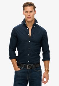 Donkerblauwe button-up shirt met een kraag, lange mouwen opgerold bij de manchetten, en een borstzak, gecombineerd met donkere spijkerbroek en een zwarte riem.