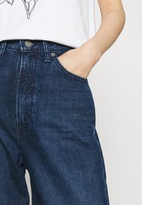 Jeans de denim azul escuro com um corte ajustado, apresentando um bolso frontal, fecho de botão metálico e uma textura subtil no tecido.