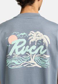 Lichtblauw T-shirt met een tekstuurafbeelding van een zonsondergang, palmbomen en oceangolven, met het woord "Ruch" in krullerige muntgroene letters.