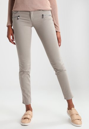 Kobieta nosząca beżowe, dopasowane spodnie z kieszeniami na suwak oraz jasnobeżowe buty wsuwane na stopę na jednolitym tle.