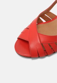Sandale en cuir rouge à bout ouvert avec des brides qui se chevauchent et un motif tissé sur un fond clair.