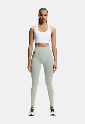 Woman with braided hair wearing a white sports bra, light green high-waisted leggings, and white sneakers, standing against a plain background.