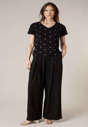 Femme debout portant un haut noir à col en V avec de petits motifs en forme de cœur, un pantalon noir à jambes larges, et des talons ouverts dorés sur un fond uni.