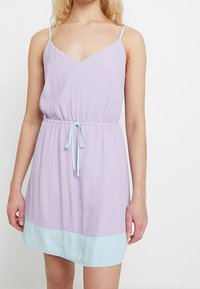 Lila sleeveless dress with a V-neck and adjustable straps. Features an elastic waistband and light blue hem panel. Smooth fabric texture.