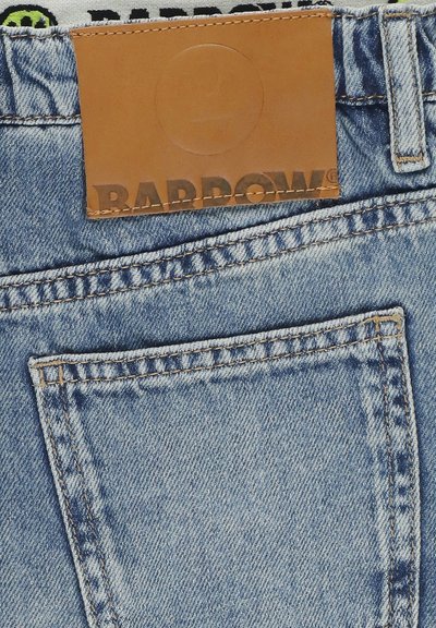 Poche arrière d'un jean en denim bleu clair avec une étiquette de marque en cuir marron cousue au-dessus, affichant partiellement le nom de la marque "BARROWS".