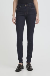 Žena v tmavě modrých úzkých džínách a černých mokasínech s třásněmi, stojící na světlém šedém pozadí.