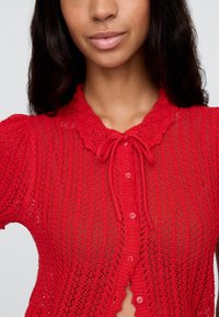 Adīts sarkans kardigans ar viļņotu apkakli, kas izceļas ar sasiešanas detaļu un pogu aizdari, demonstrējot atvērtu, mežģīņu tekstūru visā virsmā.