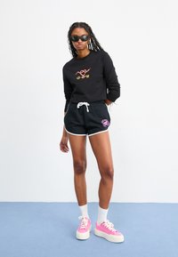 Maglietta nera a maniche lunghe con design grafico colorato, abbinata a pantaloni corti neri con rifiniture bianche e logo. Sneakers a piattaforma rosa e calze bianche.