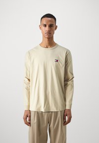 Beige, hosszú ujjú pamut póló, kerek nyakkivágással, kis, színes logóval a mellrészen, laza fazonnal.