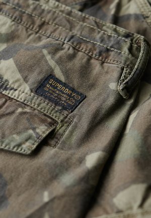 T-shirt de camouflage dans un mélange de tons verts et bruns, avec un tissu texturé, une étiquette avec "SUPERDRY" et des détails de couture.