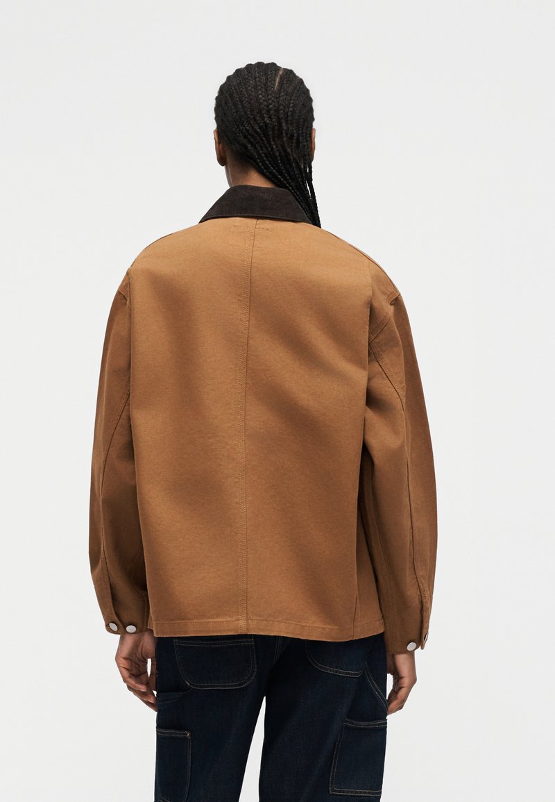 Persona con capelli intrecciati che indossa una giacca marrone con colletto scuro e pantaloni blu, vista da dietro su uno sfondo chiaro e uniforme.