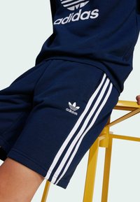 Námornícky modré šortky Adidas so zlatými tromi pásikmi po stranách a malým logom trefoil na ľavej nohe.