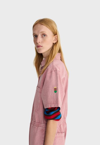 Jeune femme aux longs cheveux blonds portant une combinaison rose à manches courtes par-dessus une chemise rayée bleue, rouge et noire à manches longues, regardant de profil.