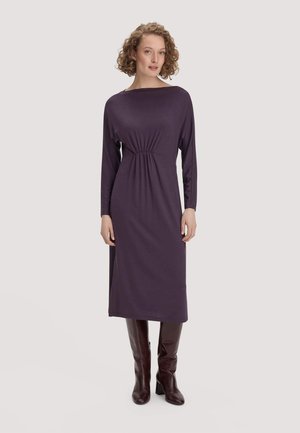 Frau mit lockigem Haar, die ein langärmeliges violettes Midikleid und kniehohe dunkelbraune Stiefel trägt, steht vor einem schlichten Hintergrund.