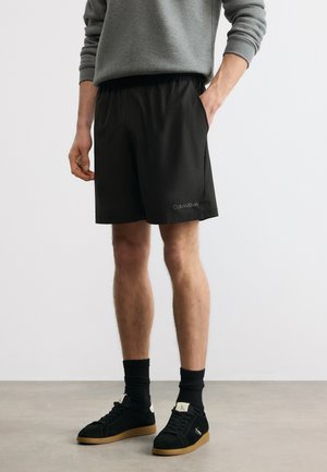 Muž oblečený v černých šortkách Calvin Klein, šedém dlouhém rukávu, černých ponožkách a černých teniskách s gumovou podrážkou stojí na šedé podlaze.