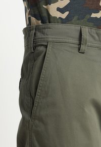Gros plan sur une personne portant un pantalon vert olive avec une poche latérale et des passants pour ceinture, associé à une chemise à motif camouflage rentrée à l'intérieur.