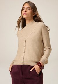 Maglione beige a maglia con collo alto e design incrociato, caratterizzato da bottoni in perla e una texture morbida. Abbinato a pantaloni in velluto a coste color prugna.