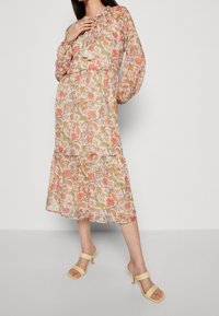 Robe midi florale en tissu léger aux teintes pastel, col froncé et manches bouffantes, avec une jupe à volants et un détail à pompons.